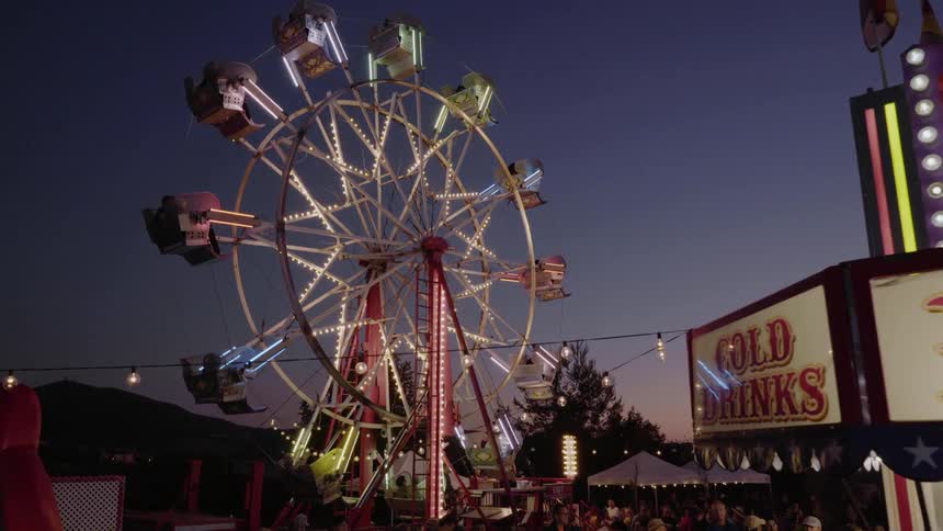 A colorful and vibrant fairground at dusk includes multiple stands for food and games as well as a brightly lit ferris wheel