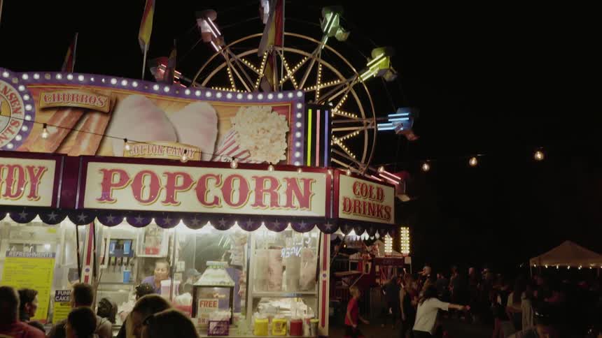 People are attending a carnival at night and buying popcorn at the food stand