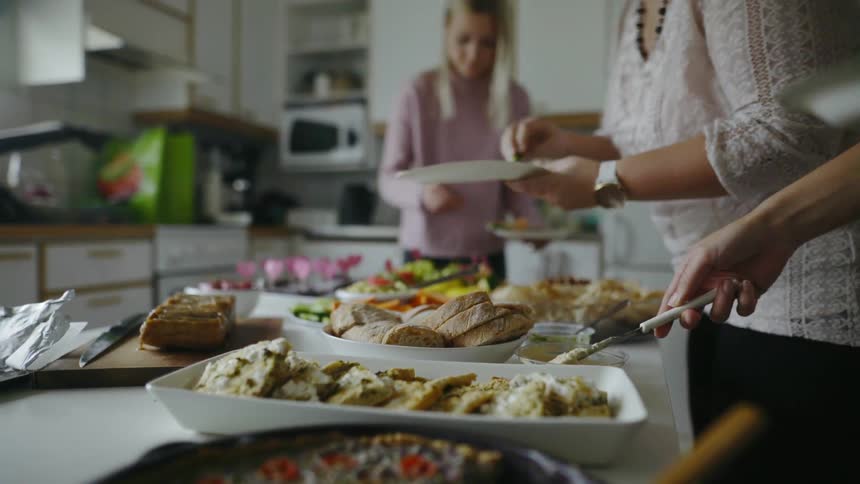 Three women put food from an assortment of dishes on a kitchen island and place it on paper plates they are holding