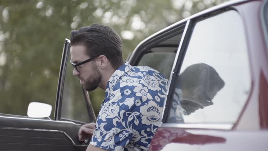 A white man wearing sunglasses gets out of a red car