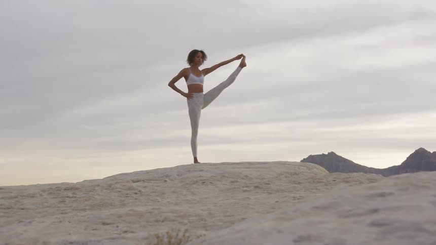A woman stretches her leg in the air standing in the dirt around mountains