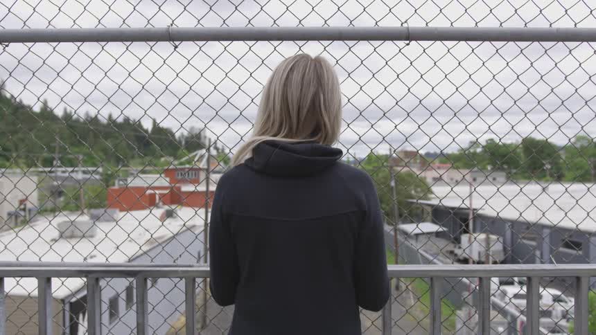 A blonde haired woman stands atop a bridge that is lined with chain linked fence and she looks out over several buildings in a forest lined town
