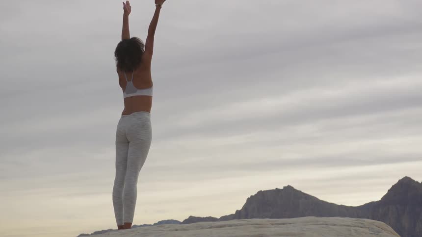 A woman standing on a cliff stretches her arms high in the air