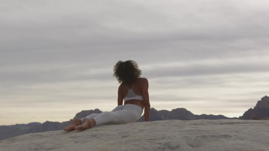A woman bends her back upwards in a yoga pose on a rocky surface outside at day