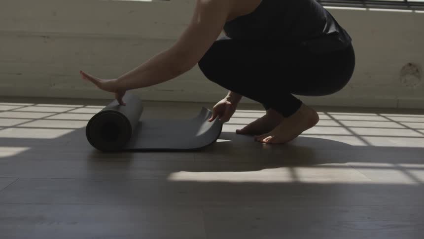 Barefoot woman rolls sports mat on the floor on a sunny day