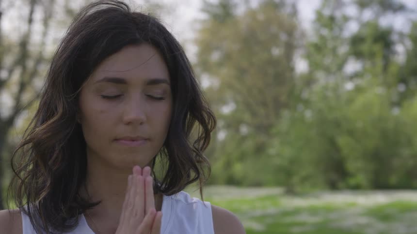 A woman is outside with her eyes closed and palms together