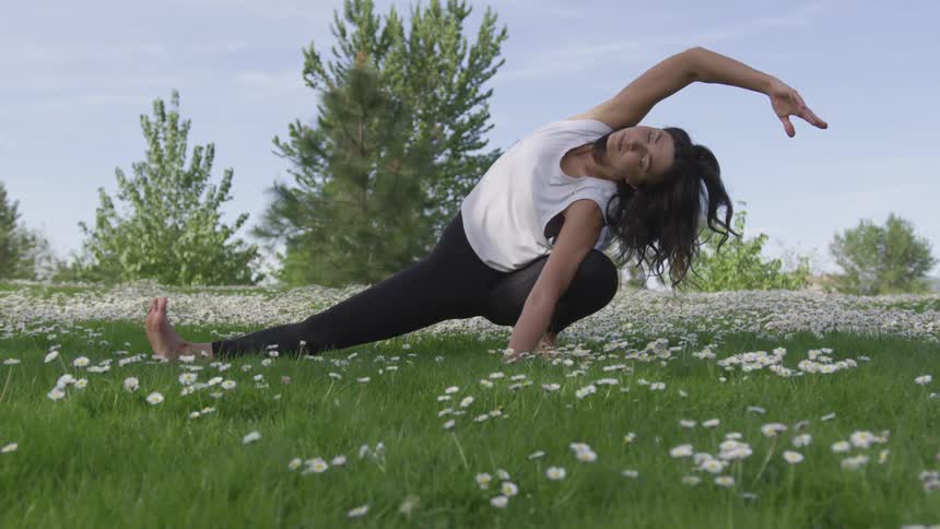 A young woman is maintaining an extreme stretching position with one leg held in a squatting position and the other outstretched as she performs her challenging yoga pose in a field full of flowers