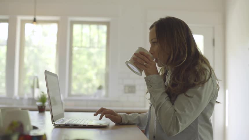 A woman is sitting at a counter top and working on her laptop while drinking from a coffee cup