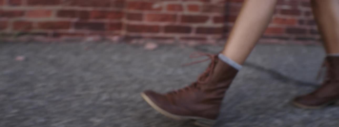 View of a woman's brown boots as she walks down a cracked, weed-filled sidewalk