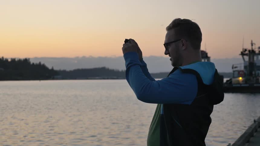 A man standing on a seawall and taking a picture with his phone