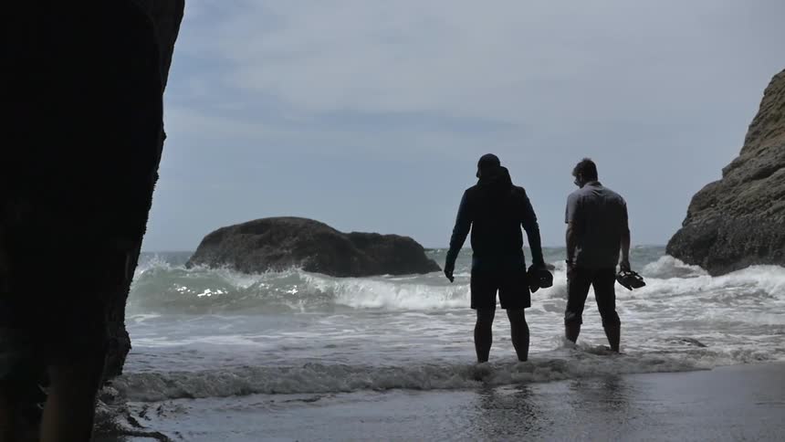 Two men carrying their shoes as they step into water that is coming on shore in an area surrounded by large boulders