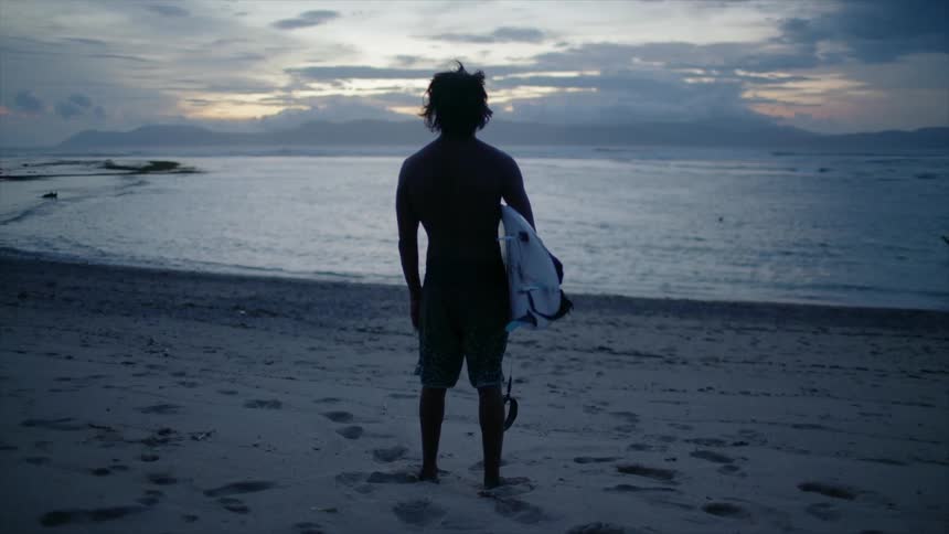 A young man holding a surfboard stands on a beach at dusk