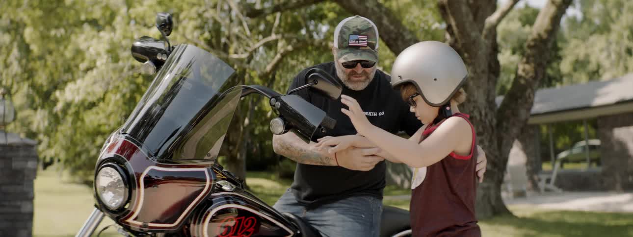 A man with a gray beard gets on a motorcycle and then helps a young boy up onto the seat of the bike in front of him