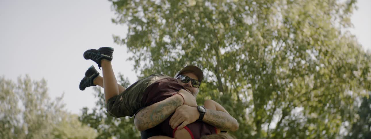 Bearded man wearing cap and sunglasses lifts up boy and plays with him in the countryside