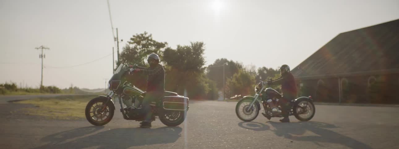 Two men each sit on their own motorcycle in a parking lot and then take off down a road