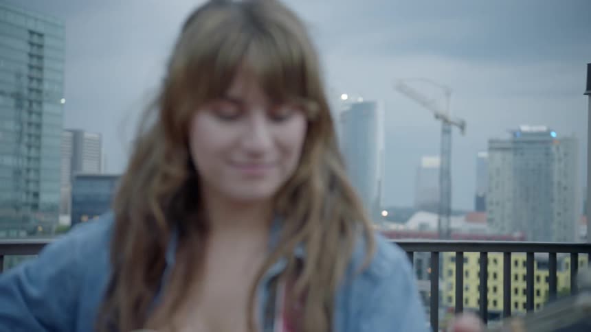 A white female plays an acoustic guitar while standing on a balcony with tall buildings and construction cranes in the distance