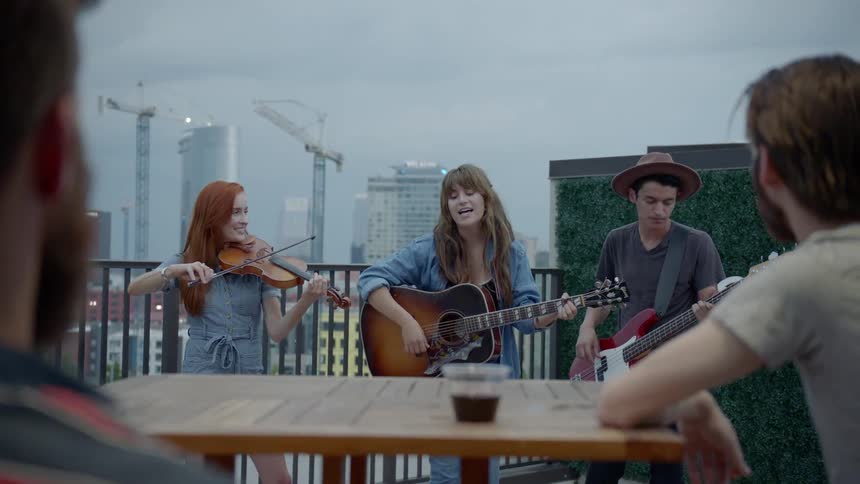 A woman strums a gibson acoustic guitar accompanied by a violinist and hat wearing bassist
