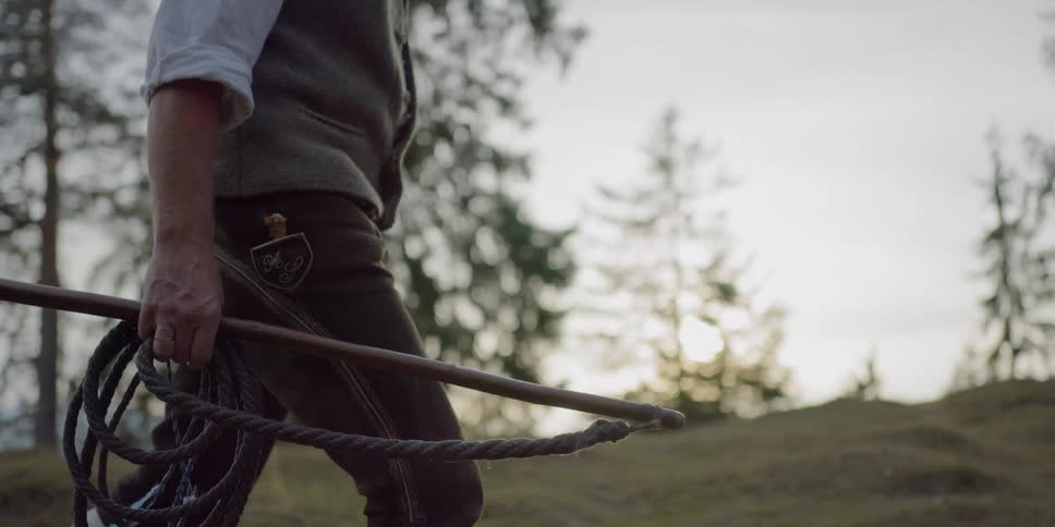 A white man walks through a field carrying a whip