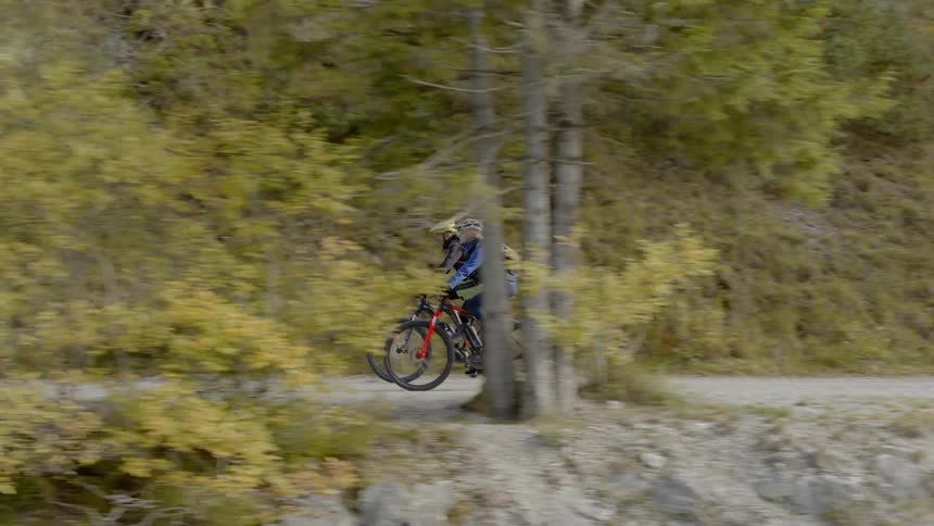 Two people bike along a path next to a river and the mountains
