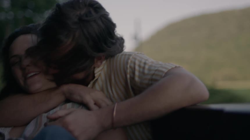 Man and woman sitting in a driving roadster car hug and laugh at sunset
