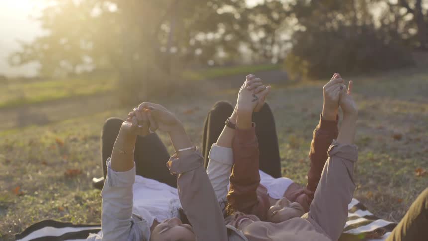 Three women lay on a blanket and raise their clasped hands