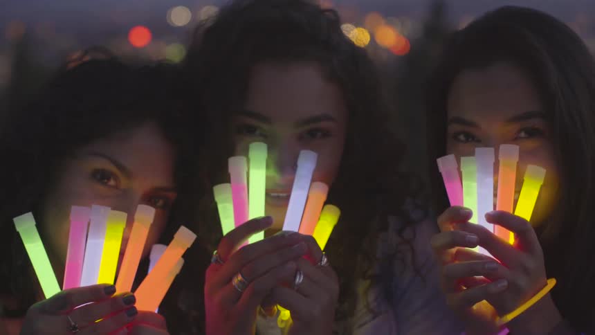 Three women hold glowing light sticks up in front of their smiling faces at night