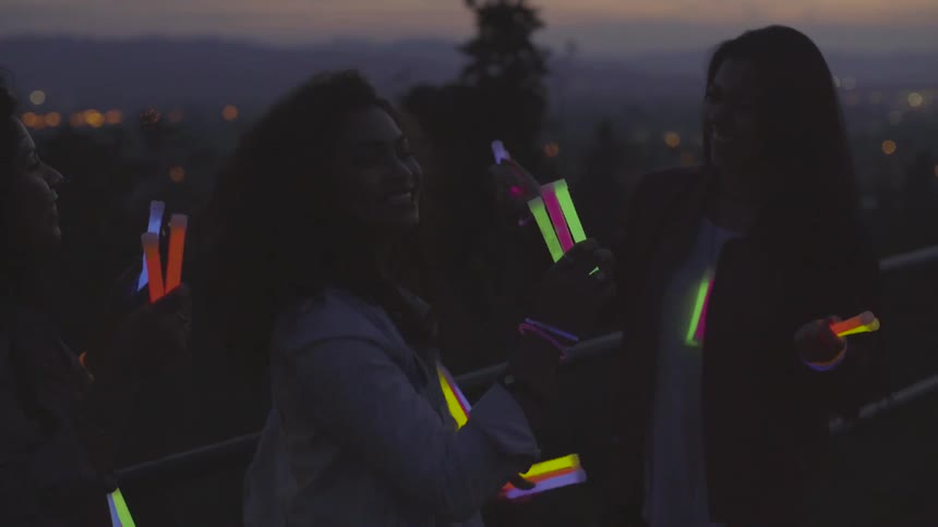 Three women dance with glow sticks outside at dusk