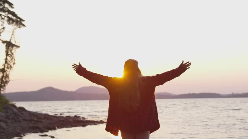A woman standing on the shore of a lake with her arms spread wide towards the rising sun