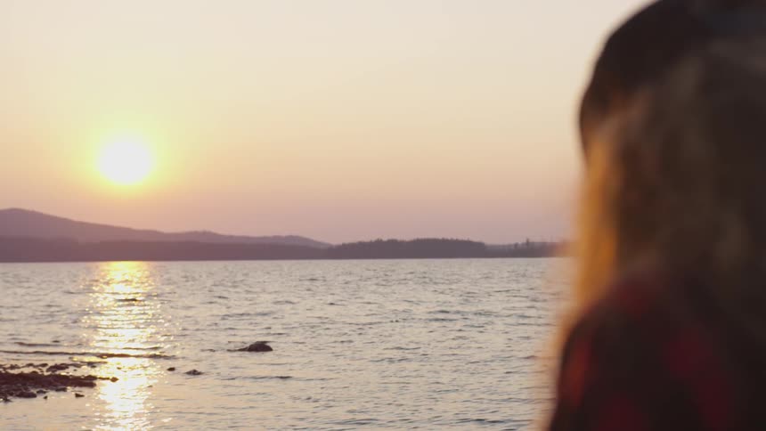 A woman with a hat staring at the lake and the sun