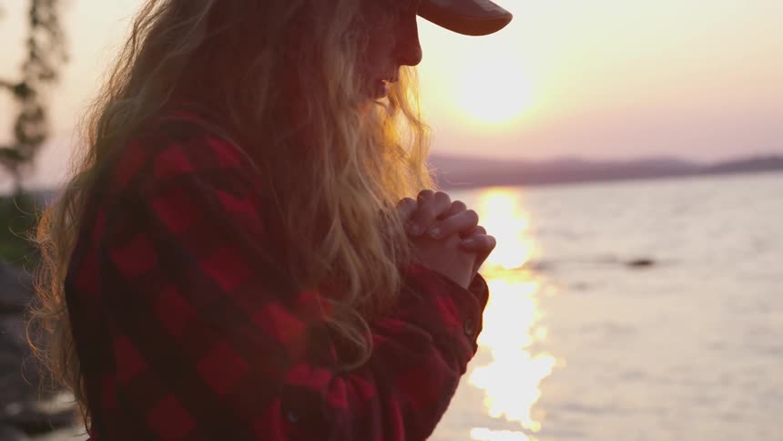 A woman stands with her hands clasped together and her head bowed overlooking the water at sunset