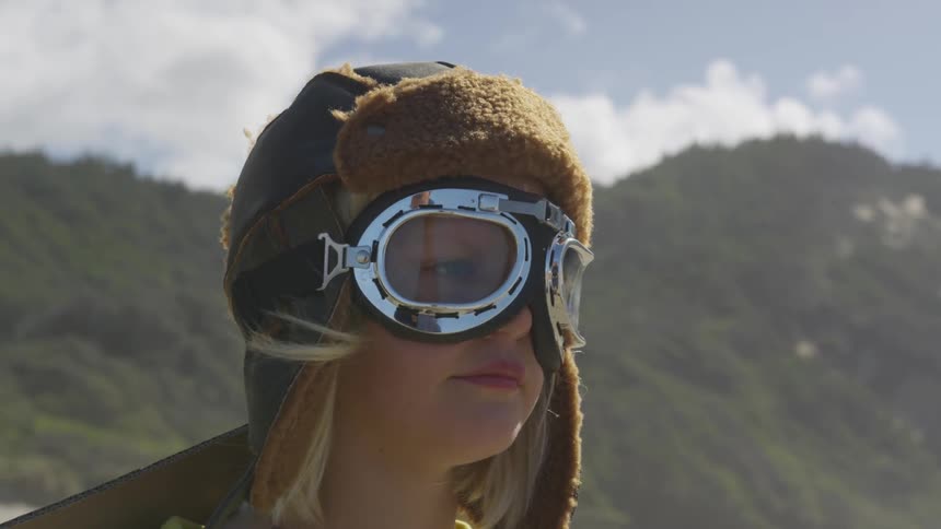 A blonde child wears an aviator hat and goggles while standing outside with a breeze blowing her hair and hills behind her