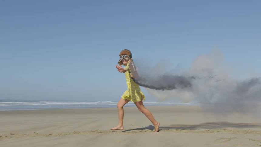 A girl in a yellow dress wears flying goggles and a helmet as well as a cape with two flares attached to look like a jetpack