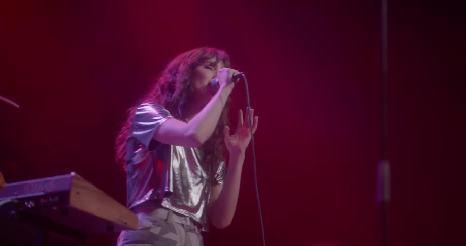 A female in a glossy crop top sings into a microphone on a stage next to a synthesizer