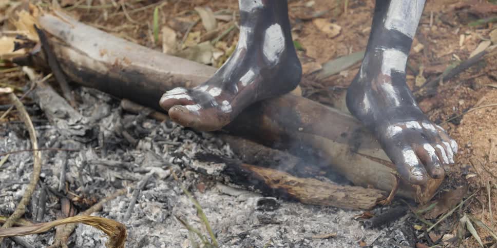 A view of an african man standing on a short log that is placed above smoking embers