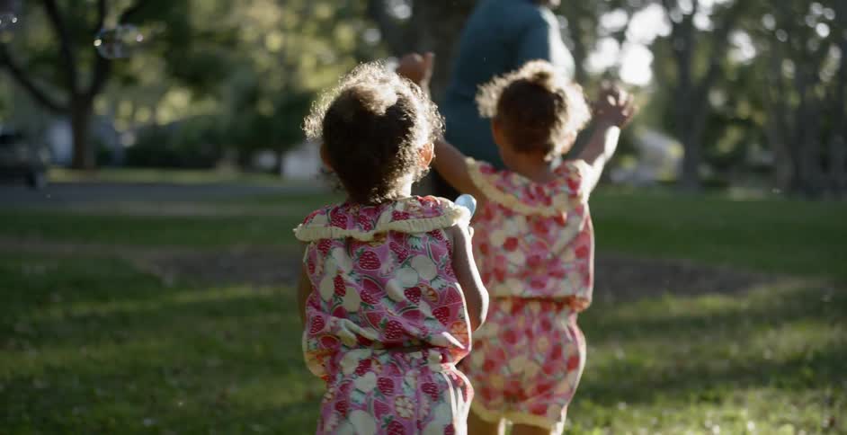 Twin little girls chasing their mom while playing at the park with bubbles