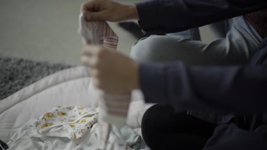 A pregnant female folding baby clothes sitting on the floor next to another person