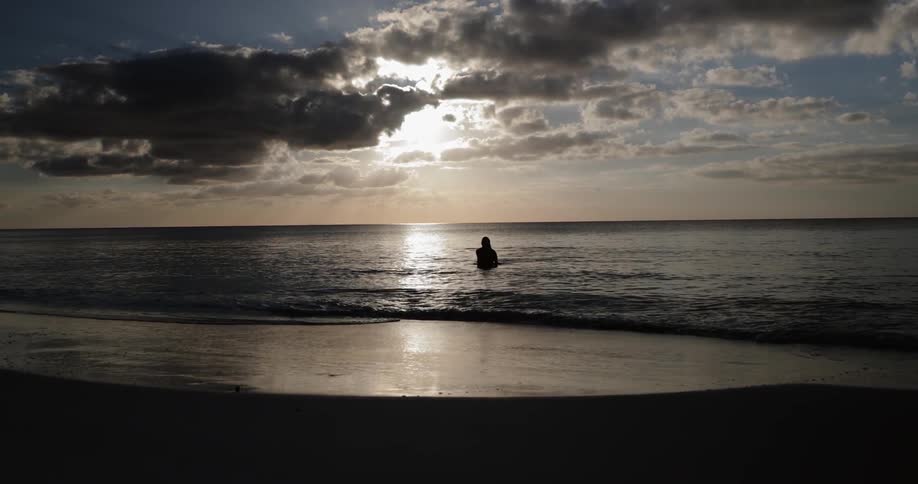 The silhouette of a person standing up and surfacing in the ocean near the sandy beach at sunset