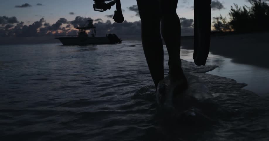 A person is walking in the water at sunset by a boat