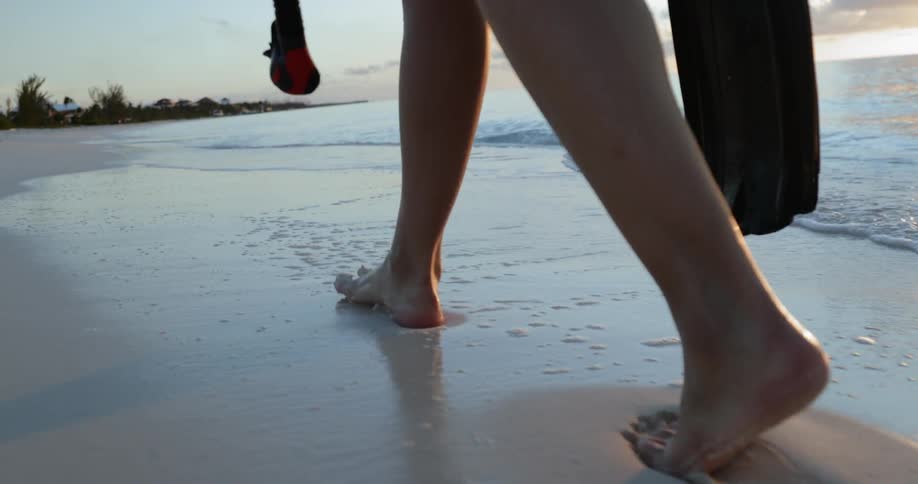 A white woman walks barefoot across a beach while holding a snorkel and flippers