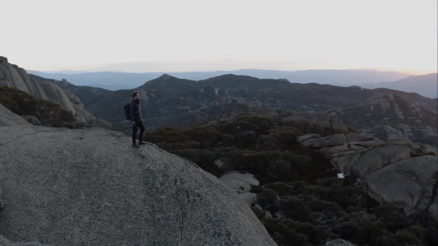 A man stands on a large mountain top looking toward the scenery