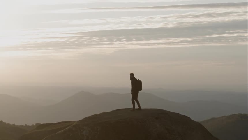A man wearing a backpack and holding a camera takes a final few steps to reach the top of hill where he stops to behold an expansive mountainous and hilly range