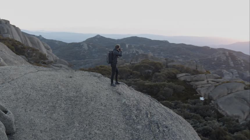 Aerial of man standing on top of some rocks at the top of a mountain taking a photo
