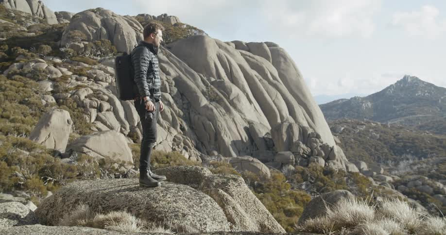 A man wearing a backpack and carrying a camera walks to a rock in a mountainous area to take a picture