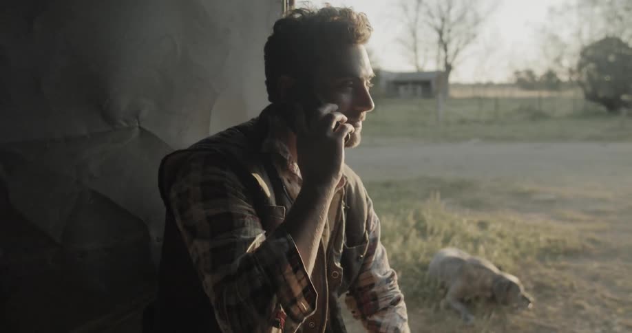 A man sitting on a stool in a doorway calls someone on his cellphone while a dog lies in the grass behind him