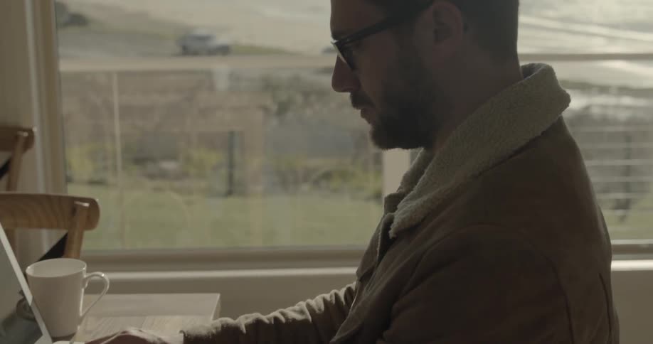 A white male sits at a table in front of a large window and types on a laptop