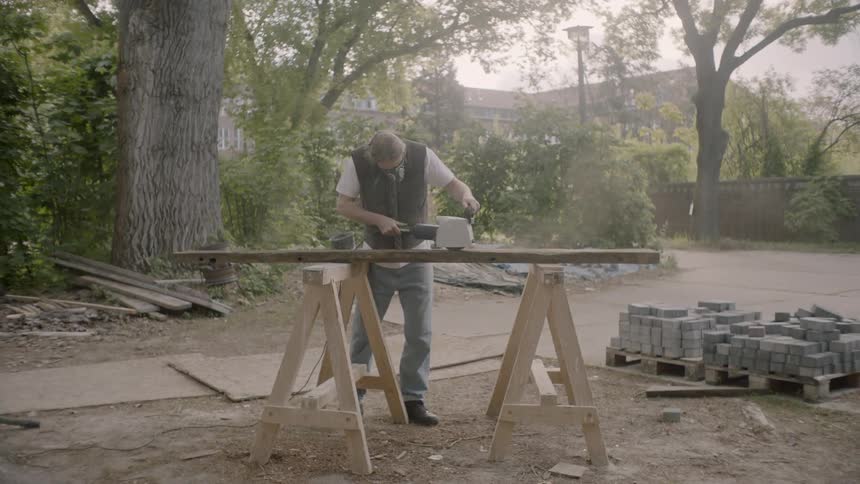 A man wearing a breathing mask sanding a piece of lumber outdoors during the day