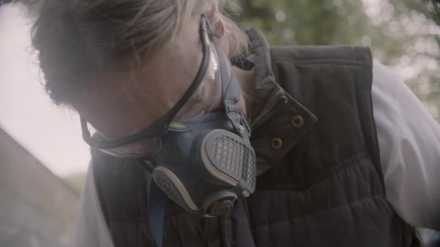 A young blonde white man with a safety mask and safety glasses sands a piece of wood