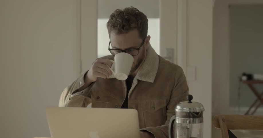Male wearing a jacket sitting in front of his laptop drinking coffee