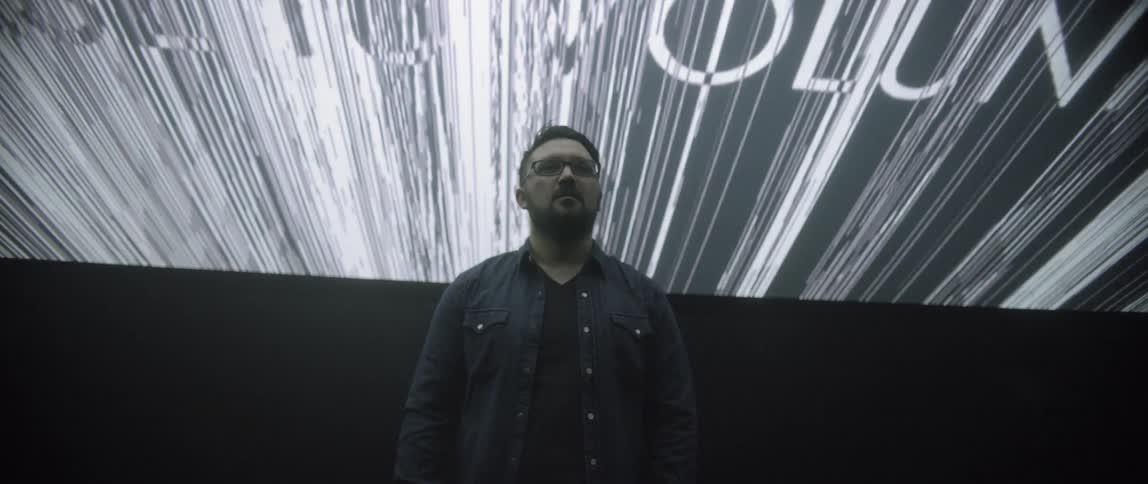 A white man stands underneath a giant video screen