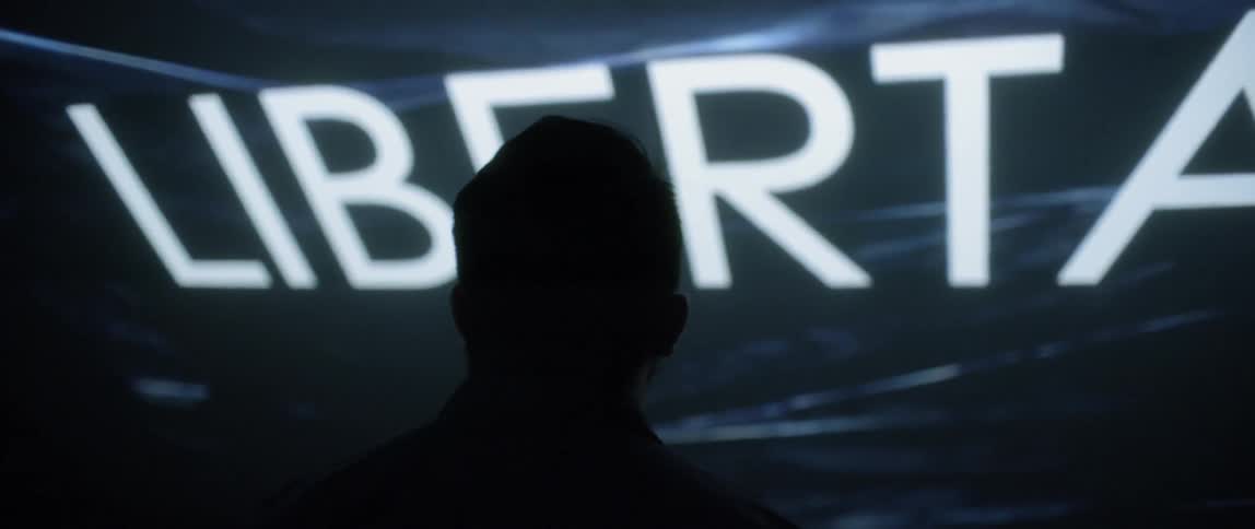A man wearing glasses looks up at a screen mounted at an angle above his head as it displays the words freedom and libertad in front of a digital background of waves